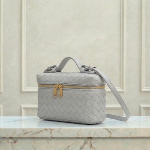 May include: A light gray woven leather vanity case with a gold zipper and hardware. The small handbag has a top handle and a detachable shoulder strap. The bag is sitting on a marble surface.