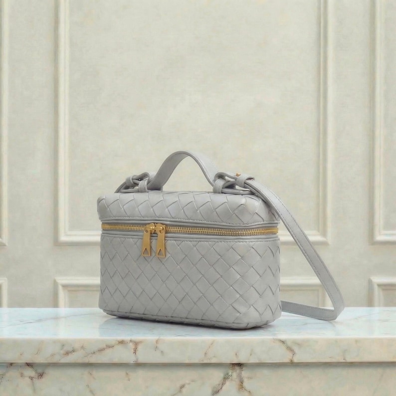 May include: A light gray woven leather vanity case with a gold zipper and hardware. The small handbag has a top handle and a detachable shoulder strap. The bag is sitting on a marble surface.