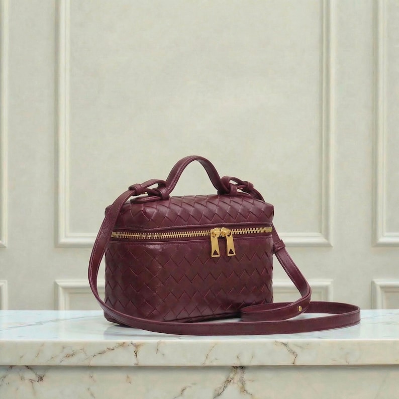 May include: A burgundy crossbody bag with a woven design. The bag has a rectangular shape with a top handle and a long, adjustable strap. It features a gold-colored zipper and hardware. The bag is sitting on a marble surface.