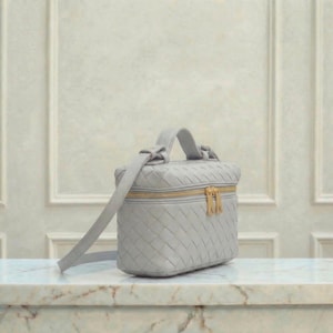 May include: A light gray, woven leather vanity case with a top handle and a long shoulder strap. The rectangular bag features a gold-colored zipper closure. The bag is sitting on a marble surface.