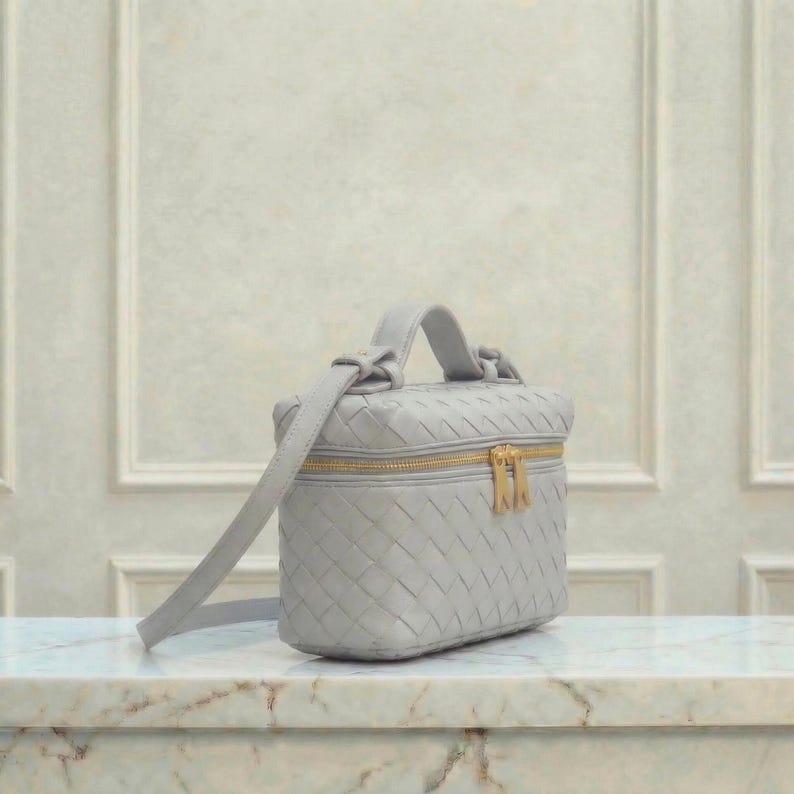 May include: A light gray, woven leather vanity case with a top handle and a long shoulder strap. The rectangular bag features a gold-colored zipper closure. The bag is sitting on a marble surface.