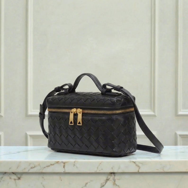 May include: Black leather crossbody bag with a woven design. The bag has a rectangular shape with a top handle and a long, adjustable strap. It features a gold-tone zipper closure and hardware.