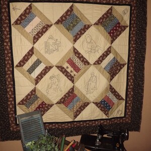 May include: A beige and brown quilt with embroidered figures of people working. The quilt is hanging on a wall with a brown floral border. Below the quilt is a sewing machine, a bucket with green plants, and a collection of wooden dowels.