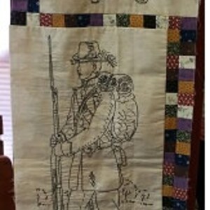 May include: Three black and white embroidered images of soldiers on a beige fabric background. The fabric is framed with a patchwork border of various colors and patterns.