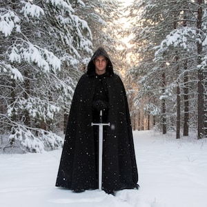 May include: A person wearing a black hooded cloak and gloves, holding a silver sword in a snowy forest. The cloak is long and flowing, and the sword is upright in the snow. The background features snow-covered trees and a bright sky.