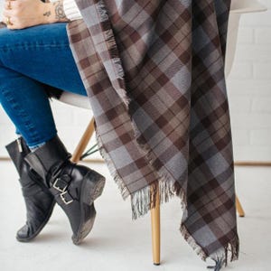 May include: A plaid scarf featuring a pattern of intersecting brown and grey lines with frayed edges. The scarf is draped over a chair, paired with blue denim jeans and black leather ankle boots that feature silver buckles on the side.