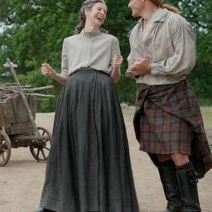 Puede incluir: Escena de un drama histórico con dos personas. Una persona con una falda larga gris oscuro y una blusa gris claro se ríe. Otra persona lleva un kilt a cuadros, una camisa clara y botas altas negras. Un carro de madera está en el fondo.