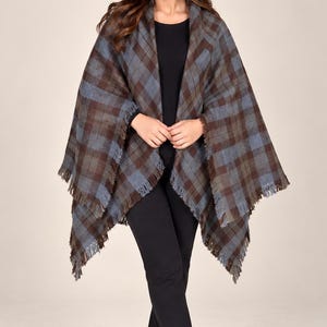 Könnte beinhalten: Ein karierter Poncho mit braun-blauem Karomuster und gefranstem Saum. Das Kleidungsstück wird über einem schwarzen Langarmshirt, einer schwarzen Hose und schwarzen Stiefeletten getragen, was einen Lagenlook mit texturierter Stoffoberfläche ergibt.