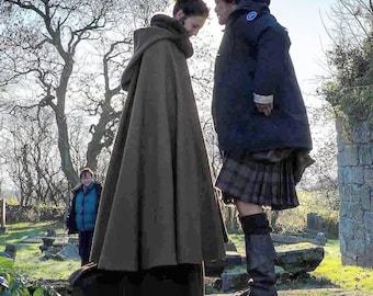 May include: Two people facing each other outdoors, one wearing a dark green hooded cloak with fur trim, the other in a navy blue coat and a plaid kilt. A third person is visible in the background, wearing a blue vest.
