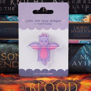 May include: A light purple and pink dragon-shaped acrylic charm on a white backing card. The card features the text Colin the cosy dragon e-reader buddy and Cosy dragon studio. The dragon has small wings, a pink belly, and a small pink bow on its head.