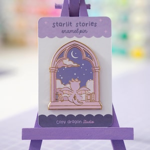 May include: An enamel pin on a backing card labeled starlit stories enamel pin and Cosy dragon Studio. The pin features a gold-outlined arched window with a purple night sky, a crescent moon, stars, clouds, and a small dragon sitting among books.
