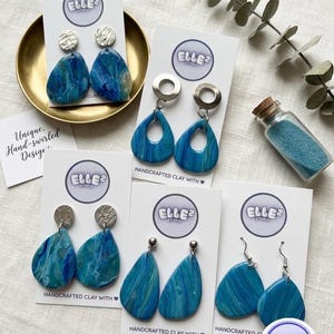 May include: Five pairs of blue marbled teardrop earrings displayed on white Elle2 Studio branded cards. The earrings feature various silver-toned hardware, including textured discs, circular hoops, and simple hooks. One pair rests in a gold bowl.