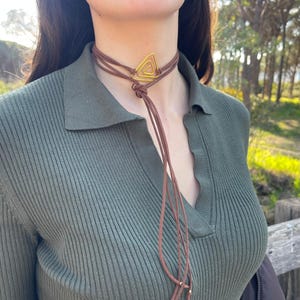 Pode incluir: Um colar gargantilha marrom com um pingente triangular dourado. O colar tem vários cordões marrons que pendem. A pessoa está usando uma camisa verde canelada com gola.