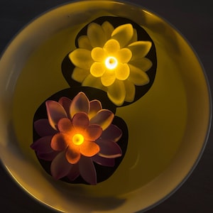 May include: Two floating, illuminated lotus flower decorations in a yellow bowl. One flower is yellow, the other is pink and orange. The flowers have a soft glow, creating a relaxing ambiance.