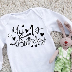 Może przedstawiać: Białe body niemowlęce z długim rękawem z czarnym napisem My 1st Birthday i małymi serduszkami. Obok body znajduje się mały pluszowy królik w zielono-białe ogrodniczki w kratkę oraz jasnoniebieski plastikowy smoczek.
