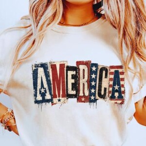 May include: White t-shirt with the word "AMERICA" in a distressed, patriotic design. The letters are in red, white, and blue, with stars and stripes. The shirt is a casual style.
