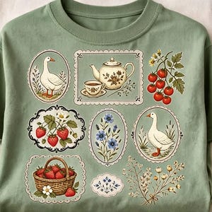 May include: Sage green t-shirt featuring framed illustrations of a goose, strawberries, a teapot, and flowers. The design includes a basket of strawberries and a sprig of tomatoes. The overall theme is a vintage, cottagecore aesthetic.