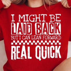 Puede incluir: Una sudadera roja con el texto blanco "I MIGHT BE LAID BACK BUT I CAN LEAN FORWARD REAL QUICK". El texto está en negrita, con una fuente desgastada. La sudadera es de color rojo liso.