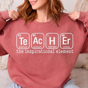 May include: A dusty rose sweatshirt with the word "TEACHER" spelled out using periodic table elements. The text "the inspirational element" is below the word teacher. The sweatshirt is a crew neck style.
