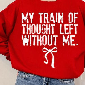 Pode incluir: Uma camisola vermelha de manga comprida com o texto branco de estilo desgastado "MY TRAIN OF THOUGHT LEFT WITHOUT ME." centrado no peito, posicionado acima de uma simples ilustração gráfica branca de um laço de fita atado.