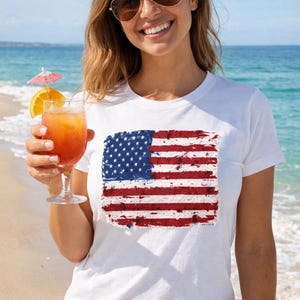 May include: White t-shirt featuring a distressed American flag design. The flag has red and white stripes and a blue field with white stars. The shirt is worn on a sunny beach day with a cocktail.