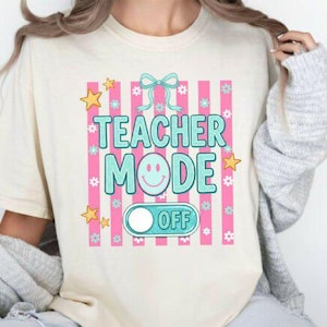 May include: Cream-colored t-shirt with a colorful graphic. The graphic features the words "TEACHER MODE" in turquoise, a smiley face, and an "OFF" switch, set against a pink striped background with stars and flowers.