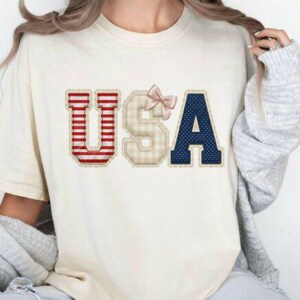 May include: Cream-colored t-shirt with the word "USA" in a decorative design. The letter "U" is red and white striped, "S" is beige plaid, and "A" is navy blue with white dots. A pink bow is above the "S".