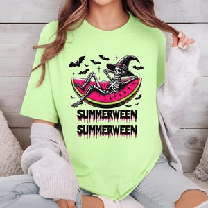 May include: A light green t-shirt with a skeleton wearing a witch hat, relaxing on a watermelon slice. The shirt has the text "SUMMERWEEN" in a spooky font, with bats in the background. A fun, seasonal graphic tee.