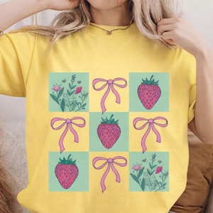 May include: Yellow short-sleeved t-shirt featuring a grid design with strawberries, bows, and floral elements. The strawberries are pink with green tops, and the bows are pink. The floral elements are green and pink.