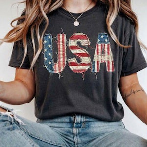Könnte beinhalten: Dunkelgraues T-Shirt mit dem Wort "USA" in einem Used-Look-Design, das an die amerikanische Flagge erinnert. Die Buchstaben sind rot, weiß und blau. Das Shirt wird mit blauen Jeans getragen.