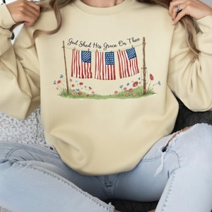 May include: Cream-colored sweatshirt featuring a graphic of three American flags hanging on a clothesline, with the text "God Shed His Grace On Thee." The flags are red, white, and blue, with a floral design at the bottom.