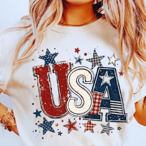 May include: Cream-colored t-shirt featuring a distressed "USA" graphic in red, white, and blue. The design includes stars in various patterns and colors, evoking a patriotic theme. The shirt has rolled-up sleeves.
