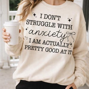 May include: A beige crewneck sweatshirt featuring black text that reads, I DON'T STRUGGLE WITH anxiety I AM ACTUALLY PRETTY GOOD AT IT. The design includes small black star icons, two small hearts, and a line-art bow illustration on the front.