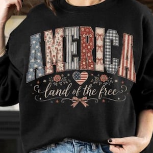 以下が含まれることがあります： 赤、白、青の模様のパッチワークデザインで「AMERICA」という言葉がプリントされた黒いスウェットシャツ。「land of the free」の文字が「AMERICA」の下にあり、ハートとリボンのグラフィックが描かれています。
