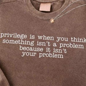 Op de afbeelding: Een bruine sweatshirt met de tekst "privilege is wanneer je denkt dat iets geen probleem is omdat het niet jouw probleem is" in het wit. Een gouden hangerketting is zichtbaar.