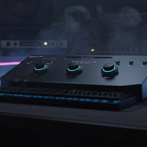 May include: Black audio mixing console with three large knobs and a small keyboard. The word "FUSION" is visible on the console. The console has blue and teal accent lighting. The background includes speakers and other audio equipment.