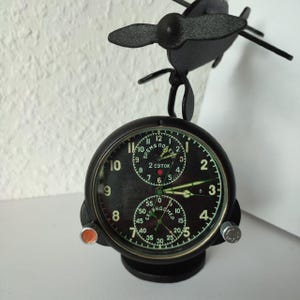 May include: A black metal aviation-style clock featuring a propeller top ornament. The round face has a black background with white numerals, two sub-dials, and Cyrillic text. It includes a red button on the left and a silver knob on the right side.