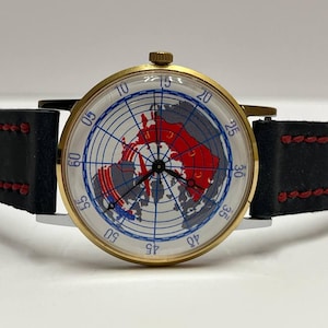 May include: A gold-tone wristwatch with a white dial featuring a blue grid map of the northern hemisphere, red landmasses, and the Cyrillic text CCCP. The watch has black hands and a black leather strap with red stitching and a gold-tone crown.