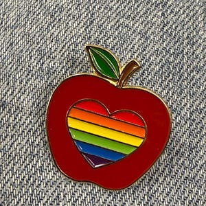 May include: A metal enamel pin shaped like a red apple with a green leaf. The center of the apple features a heart-shaped cutout filled with horizontal stripes in rainbow colors including red, orange, yellow, green, blue, and purple.