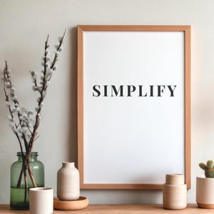 Puede incluir: Una impresión enmarcada con la palabra "SIMPLIFY" en letras negras. La impresión está en un marco de madera clara y se exhibe sobre una superficie de madera con jarrones decorativos, una botella de vidrio con ramas y un pequeño cactus en maceta.