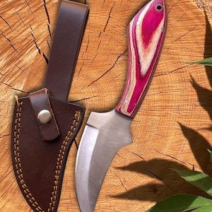 Può includere: Un piccolo coltello curvo con un manico in legno rosa e bianco e una lama in acciaio inossidabile. Il coltello è abbinato a un fodero in pelle marrone con passante per cintura. Il fodero è cucito con un filo a contrasto e fissato con un bottone automatico in metallo.