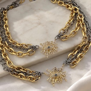 May include: A multi-strand layered necklace featuring alternating gold-tone and dark gunmetal-tone chain links. Two sunburst-shaped pendants with clear baguette-cut crystals and a central dark blue stone accent are attached to the chains.