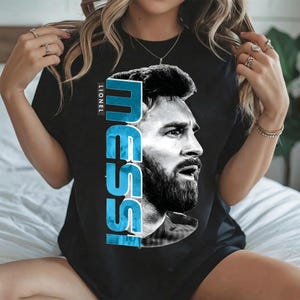 Puede incluir: Una camiseta negra de manga corta con un gran estampado gr&aacute;fico de Lionel Messi de perfil. El dise&ntilde;o incluye el nombre MESSI en letras may&uacute;sculas verticales de color azul claro y el nombre LIONEL impreso en blanco m&aacute;s peque&ntilde;o arriba.