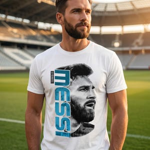 Puede incluir: Una camiseta blanca de manga corta con un retrato en blanco y negro de Lionel Messi. El nombre "MESSI" est&aacute; impreso verticalmente en grandes letras azules junto al retrato, con la palabra "LIONEL" escrita en texto m&aacute;s peque&ntilde;o arriba.