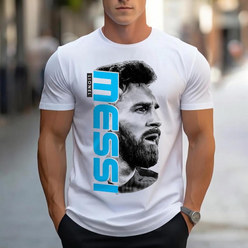 Puede incluir: Una camiseta blanca de manga corta con un retrato fotogr&aacute;fico en blanco y negro de Lionel Messi en la parte delantera. El nombre MESSI est&aacute; impreso verticalmente en letras may&uacute;sculas grandes, negritas y azul claro junto al retrato, con LIONEL encima.