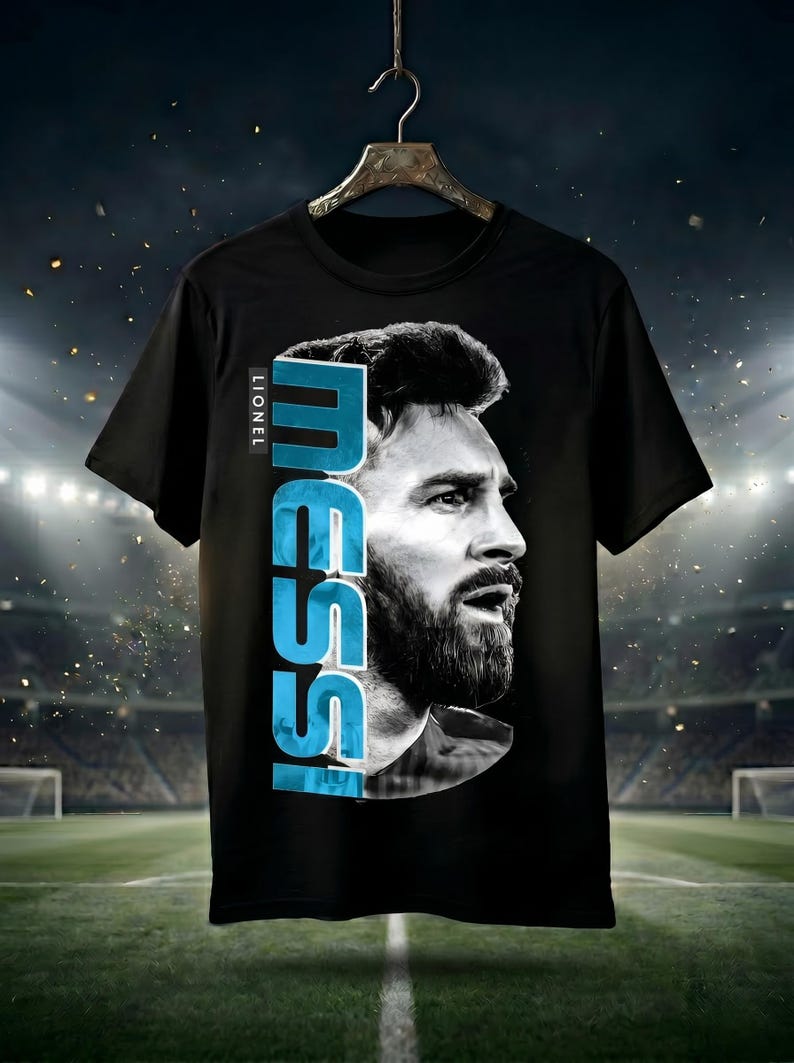 Puede incluir: Una camiseta negra de manga corta con un gran retrato en blanco y negro de Lionel Messi de perfil. El nombre MESSI est&aacute; impreso verticalmente en letras may&uacute;sculas de color azul claro junto al retrato, con la palabra LIONEL encima.