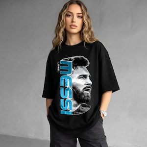 Puede incluir: Una camiseta negra de manga corta con un estampado gr&aacute;fico de Lionel Messi. El dise&ntilde;o incluye un retrato en blanco y negro del atleta junto al texto azul vertical "MESSI" y el texto blanco m&aacute;s peque&ntilde;o "LIONEL" en el lado izquierdo.