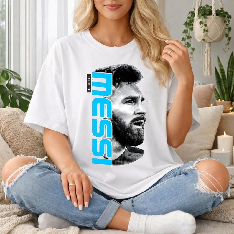 Puede incluir: Una camiseta blanca de manga corta con un retrato en blanco y negro de Lionel Messi en la parte delantera. El nombre MESSI est&aacute; impreso verticalmente en letras may&uacute;sculas grandes, negritas y azul brillante junto al retrato, con LIONEL arriba.