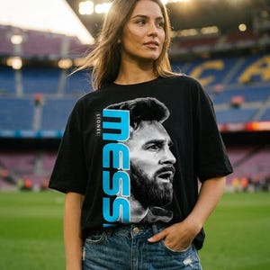 Puede incluir: Una camiseta negra de manga corta que presenta un gran retrato en blanco y negro de Lionel Messi. El nombre MESSI est&aacute; impreso verticalmente en letras may&uacute;sculas de color azul claro junto al retrato, con el nombre LIONEL en texto blanco.