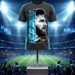 Puede incluir: Una camiseta de manga corta de color gris carb&oacute;n que presenta un retrato de perfil en blanco y negro de Lionel Messi. El nombre MESSI est&aacute; impreso verticalmente en letras may&uacute;sculas grandes de color azul brillante junto al retrato, con el nombre LIONEL en texto m&aacute;s peque&ntilde;o.